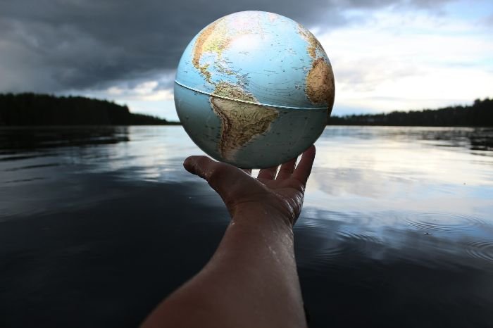A hand holding a globe against a dramatic sky over a tranquil lake, symbolizing the significance of understanding global climate zones for effective environmental management and gardening.