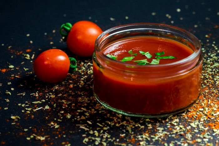 A glass jar of rich, freshly made tomato sauce garnished with parsley, with whole ripe tomatoes and a sprinkle of herbs in the background, symbolizing the tomato's vital role in global cuisine.