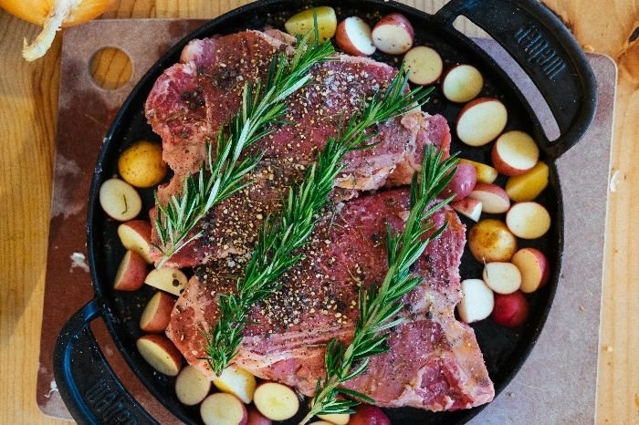 Fresh rosemary sprigs rest atop a generously seasoned steak surrounded by a medley of multicolored potatoes, ready for roasting, showcasing the herb's essential role in enhancing culinary dishes.
