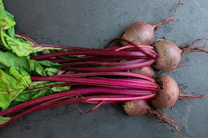 Freshly harvested beets with long, crimson stems and vibrant green leaves on a dark grey background, emphasizing their cardiovascular health benefits.