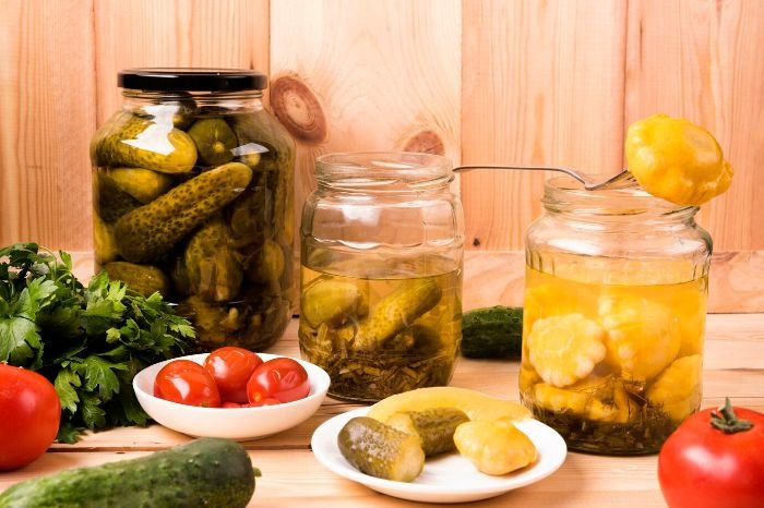 Jars of pickled vegetables with fresh tomatoes, cucumbers, and herbs on a wooden table, illustrating the steps of peeling and preparing veggies for canning.