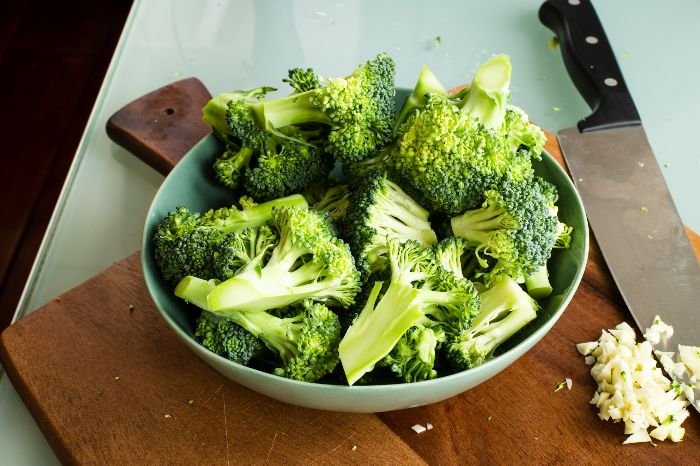 Fresh broccoli florets in a teal bowl on a wooden cutting board, next to chopped garlic, ready for cooking, highlighting the ease of incorporating broccoli into daily meals.