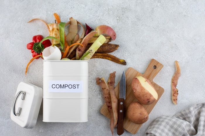 A compost bin filled with kitchen scraps like vegetable peels and eggshells, next to a cutting board with potatoes and a knife, illustrating the initial steps of setting up a compost bin for turning waste into nutrient-rich soil.