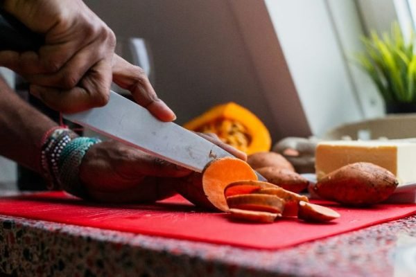 Close-up of hands skillfully slicing sweet potatoes on a vibrant red cutting board, illustrating the preparation of this vitamin-rich vegetable for healthy meals.