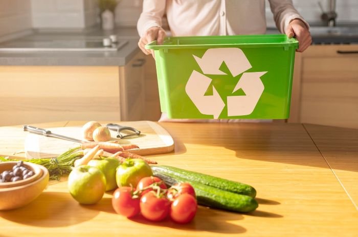 A person holding a green recycling bin in a kitchen, with fresh vegetables on the counter, illustrating the use of organic matter like compost to lower soil pH for azaleas.
