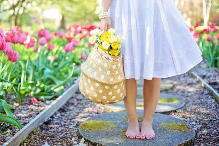 Woman in a white dress holding a basket of fresh flowers, walking through a biodynamic garden with blooming tulips.