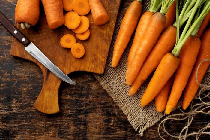 Fresh carrots on a wooden table, with sliced pieces on a cutting board, highlighting their beta-carotene content that protects the skin from aging and damage.