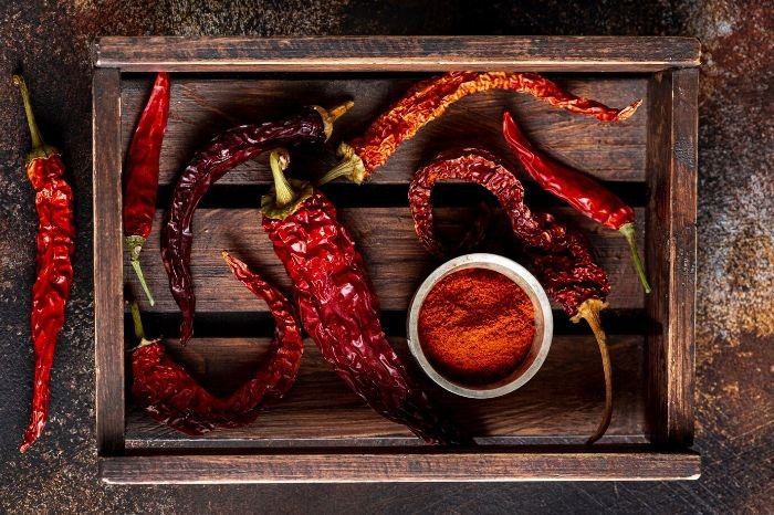 Fresh and dried chili peppers in a wooden crate with a bowl of chili powder, showcasing their versatile uses in stir-fries, soups, salads, sauces, and marinades for added spice and flavor.
