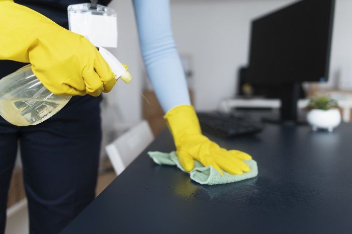 Person in yellow gloves cleaning a desk with a spray bottle and cloth, showcasing efficient cleaning techniques for busy lifestyles.