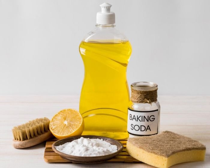 Essential ingredients for DIY natural cleaners, including a bottle of yellow liquid soap, a jar labeled "baking soda," a half lemon, a wooden brush, and a sponge, arranged on a light surface.