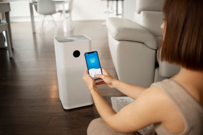 Woman using a smartphone to control a smart air purifier in a modern living room, demonstrating how technology enhances indoor air quality and comfort.