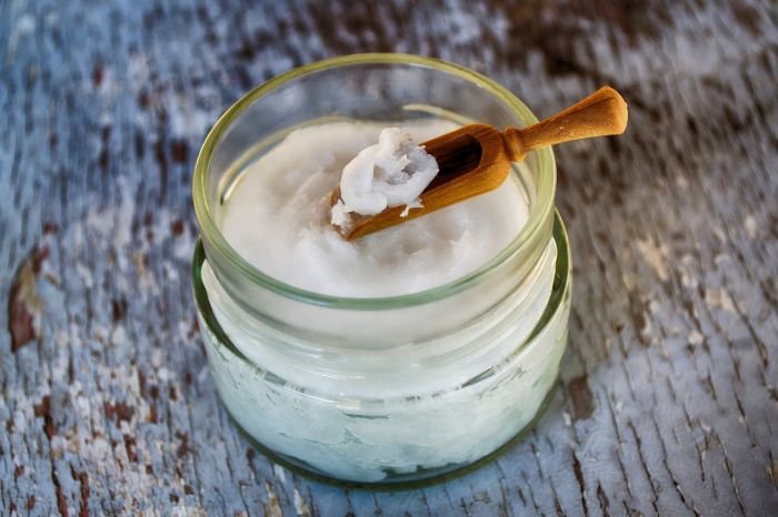 A jar of coconut oil with a small wooden scoop resting on top, showcasing a natural and non-toxic option for polishing wood furniture, placed on a rustic wooden surface.