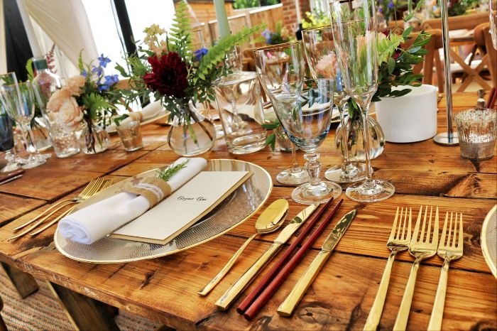 A rustic dinner table adorned with elegant gold cutlery, glassware, and floral centerpieces, featuring a personalized menu and thank-you note wrapped in a linen napkin for a thoughtful, unique touch.