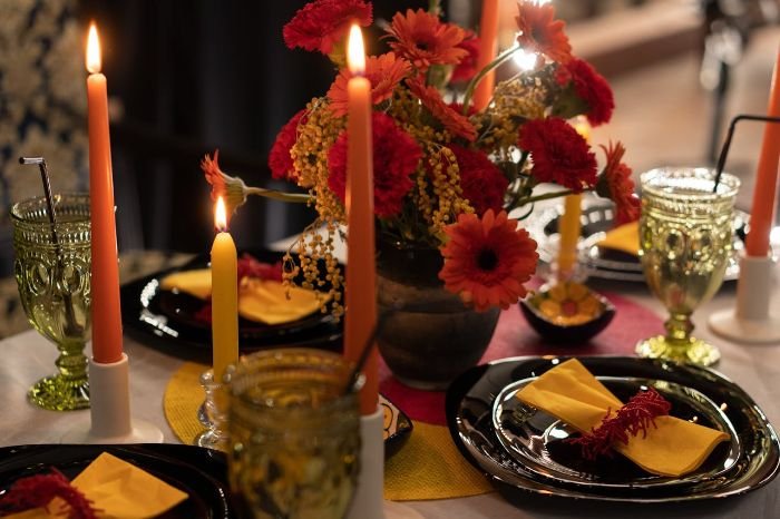 A beautifully set dinner table featuring vibrant red flowers as a centerpiece, glowing candles, elegant glassware, and personalized place settings with yellow napkins for a thoughtful and welcoming touch.