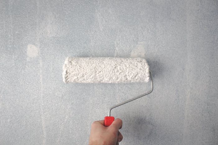 A hand holding a paint roller applying white paint on a repaired drywall surface, demonstrating techniques for blending and painting to achieve a seamless finish.