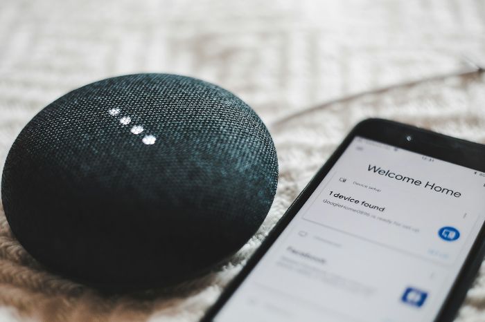 A smart speaker and smartphone displaying a smart home app interface, illustrating Wi-Fi, Bluetooth, and smart hub integration for connected devices like smart thermostats.