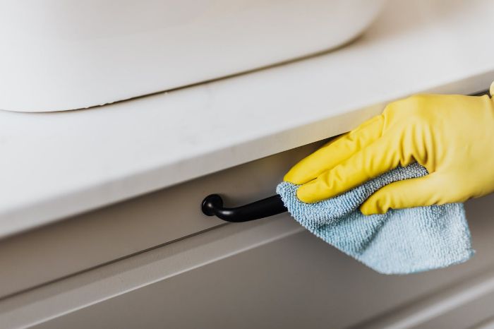 Gloved hand using a blue microfiber cloth to clean a drawer handle, demonstrating a reusable and eco-friendly cleaning alternative to paper towels.